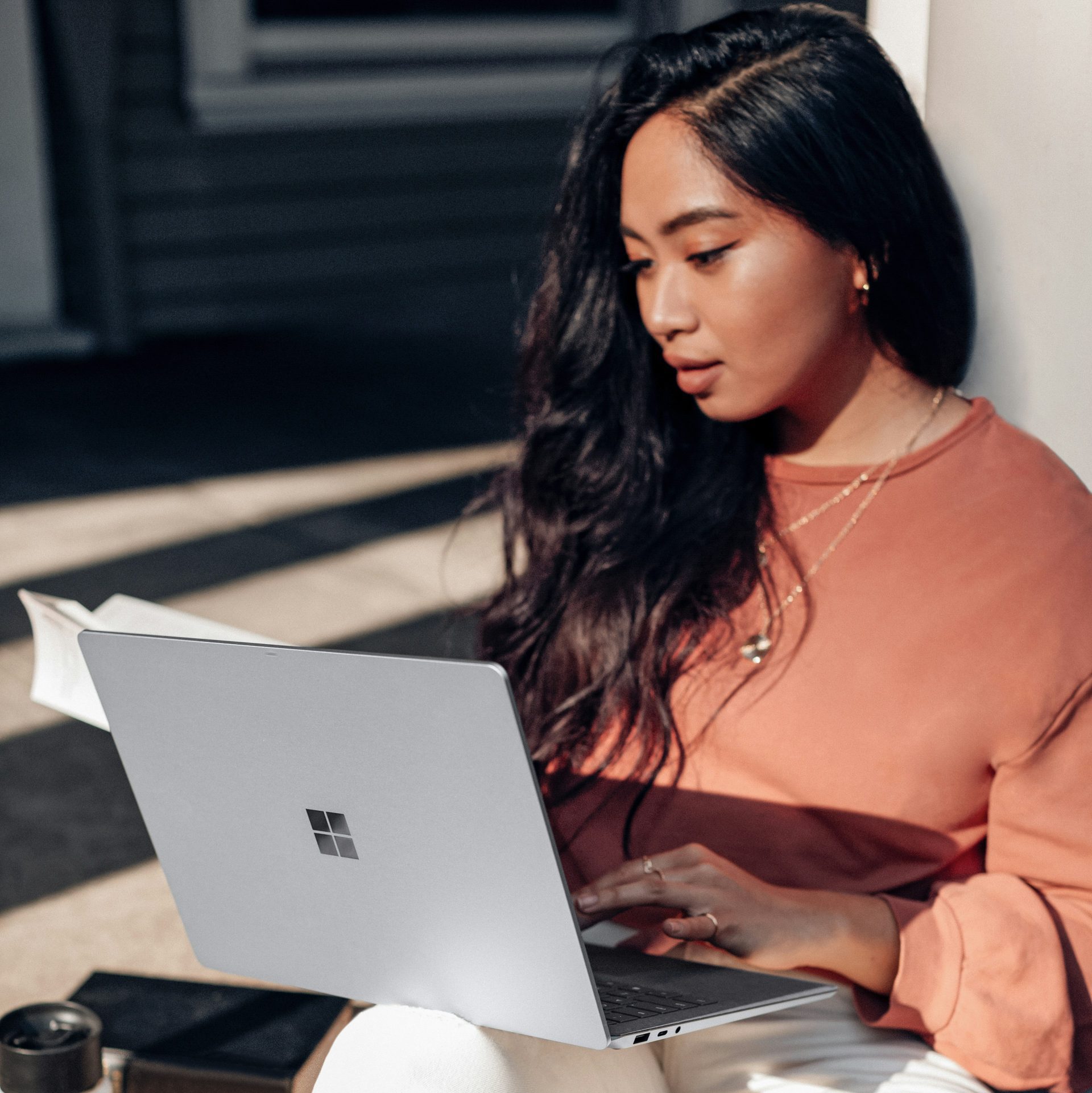 woman in orange long sleeve shirt and white pants sitting on floor using Surface Laptop